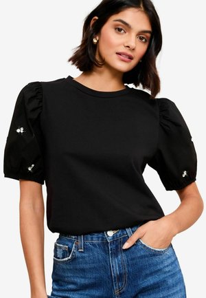 Top nero a maniche corte con maniche a palloncino e polsini elastici, impreziosito da piccoli dettagli ricamati, abbinato a jeans in denim blu.