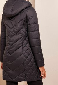 Žena v černé prošívané bundě s kapucí, zdobené vzory v kosočtvercích a šipek, stojí u světle béžové zdi.