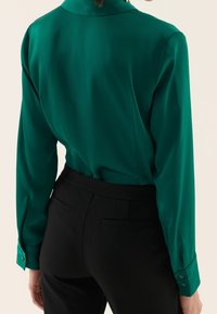 Blusa verde lucida con maniche lunghe e polsini abbottonati, abbinata a pantaloni neri su misura con tasche posteriori e tessuto liscio.