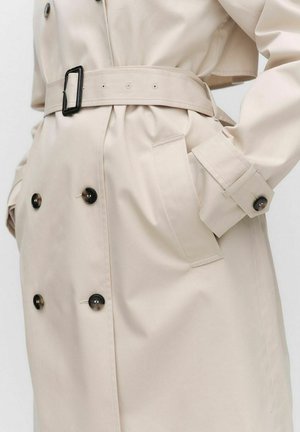 Beige trench coat, jossa on kaksiriviset napit, vyö vyötäröllä ja kaksi sivutaskua. Sujuva tekstuuri ja muotoiltu istuvuus.
