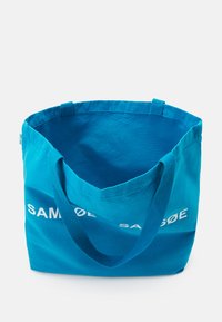 Μπλε βαμβακερή τσάντα tote με ανθεκτικά χερούλια. Ο εσωτερικός χώρος είναι ευρύχωρος. Στο μπροστινό μέρος υπάρχει το όνομα της μάρκας "SAMSØE" σε λευκό χρώμα.