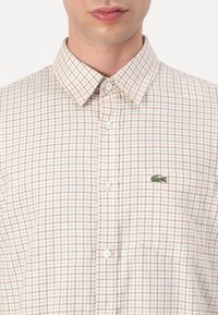 Button-up shirt in light plaid pattern of beige and brown. Features a collared neckline and a green embroidered logo on the chest pocket.
