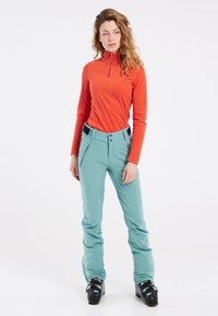 Oranges Langarm-Top mit Viertel-Reißverschluss, kombiniert mit hellblauen Skihosen, die über Reißverschlusstaschen und eine körperbetonte Passform verfügen. Schwarze Skistiefel runden das Outfit ab.