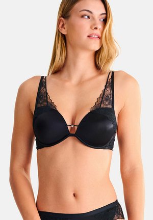 Femme portant un soutien-gorge noir bordé de dentelle et un sous-vêtement assorti, regardant vers sa gauche sur un fond uni.