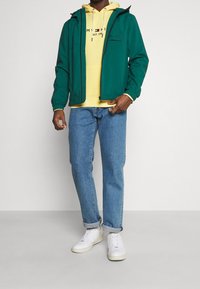Chaqueta verde con cremallera y capucha sobre una sudadera amarilla, combinada con jeans de color azul claro y zapatillas blancas. Presenta detalles del logo en la sudadera.