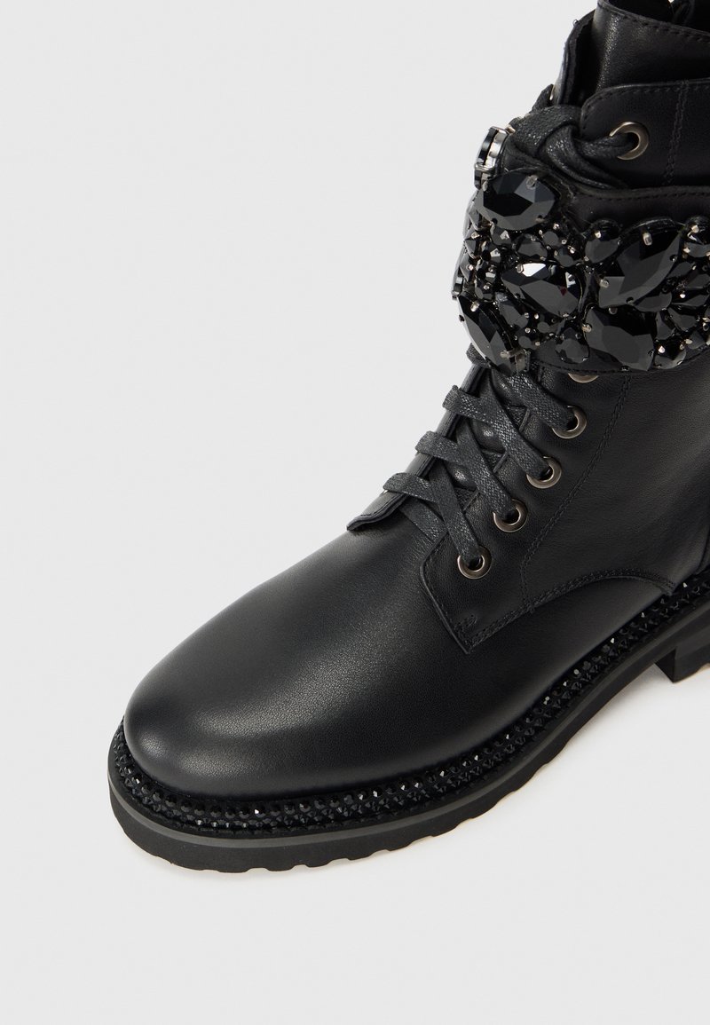 Stivale alla caviglia in pelle nera con tomaia testurizzata, allacciato sul davanti e con accentuazioni di gemme nere decorative attorno al polsino. Suola spessa e con battistrada.