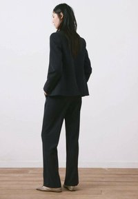 Mujer de pie con las manos en los bolsillos, vestida con un traje negro a rayas y zapatos planos beige, mirando hacia la izquierda contra una pared blanca lisa.