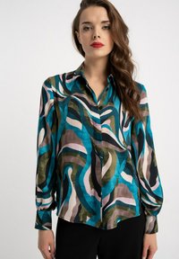 Kobieta ubrana w koszulę na guziki z długim rękawem o abstrakcyjnym wzorze z turkusowych, zielonych, brązowych, czarnych i różowych zawirowań, połączoną z czarnymi spodniami.