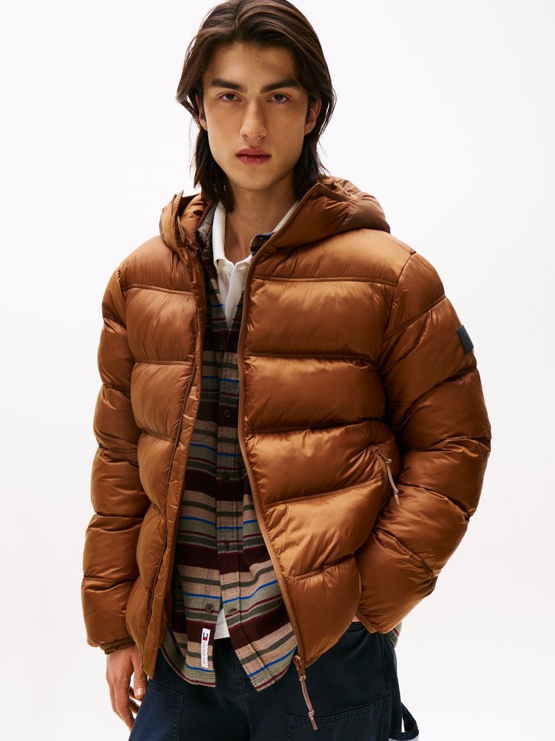 Giacca a vento marrone con cappuccio, design trapuntato e tasche con zip, indossata sopra una camicia tartan multicolore. Tessuto liscio e lucido.