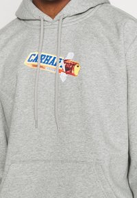 Grijze hoodie van een katoenmix, met een kleurrafisch van een snoepwikkel waarop de tekst "CARHARTT" staat, en zilveren koordaccenten.
