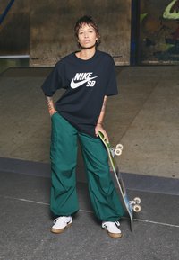 Black Nike SB t-shirt, green baggy pants, and white sneakers with a gum sole. Holding a skateboard, standing on a textured surface.
