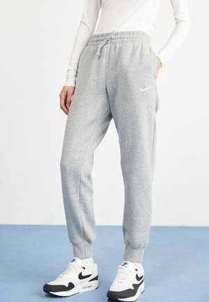 Persona que lleva pantalones deportivos Nike gris claro con puños elásticos y zapatillas Nike blancas, de pie sobre suelo azul frente a un fondo blanco.