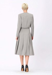 Robe grise sur mesure avec des épaules structurées, jupe plissée et taille cintrée. Portée avec des talons hauts noirs, offrant une texture de tissu lisse.