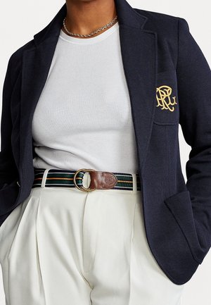 Personne portant un blazer bleu marine avec un logo doré, une chemise blanche côtelée, un pantalon plissé crème, une ceinture rayée avec une boucle en cuir et un collier chaîne argenté.