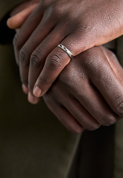 Bague en argent avec un design texturé, portée sur une main à la peau foncée, entrelacée avec une autre main, mettant en valeur une peau lisse et une couleur de vernis à ongles naturelle.