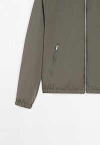 Light brown jacket with front zipper, zippered side pocket, and elastic cuffs on sleeves and hem.