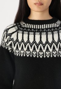 Schwarzer Pullover mit einem weißen geometrischen Muster entlang des Halsausschnitts. Das Material sieht weich und strukturiert aus und hat einen runden Halsausschnitt.