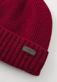 Piros kötött sapka bordás textúrával, amelyen egy varrott barna bőrcímke található, rajta "Barbour England" felirattal.