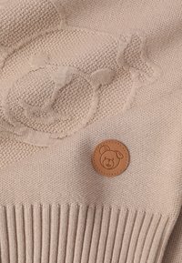 Tessuto lavorato a maglia beige con un motivo testurizzato di orso e una toppa in pelle rotonda con il logo di un orso. Dettagli a costine lungo il bordo inferiore.