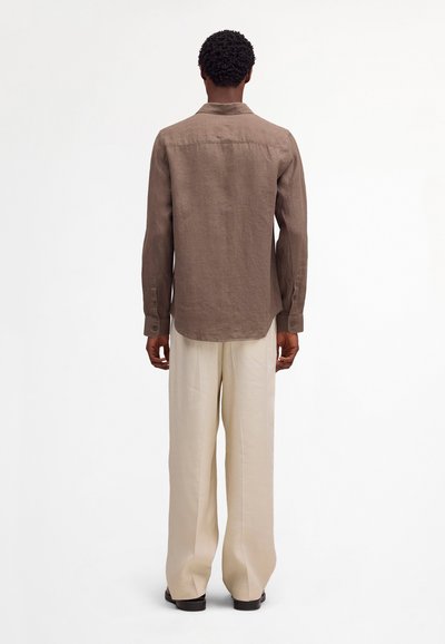 Homme portant une chemise marron à manches longues et un pantalon large beige clair, debout de dos sur un fond blanc uni.