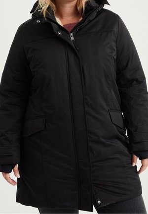 Manteau d'hiver noir matelassé avec fermeture éclair à l'avant et boutons-pression, poches à rabat, et pull marron visible en dessous, porté par une personne.