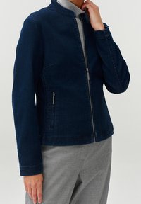 Person som bär en mörkblå zip-up-jacka med dragkedjefickor över en ljusgrå polotröja och grå byxor, hand som rör vid halsen.