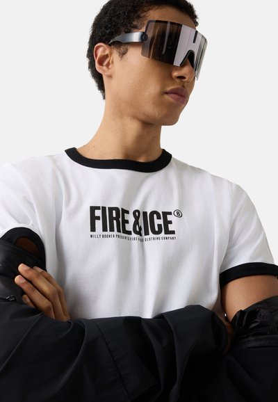 Mand iført sort-hvid "FIRE & ICE" t-shirt og store sorte skygge solbriller, med armene krydset og holder en sort jakke.
