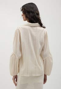 Woman with long curly hair wearing a cream-colored, striped blouse with puffed sleeves, turned away against a plain background.