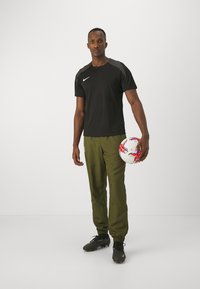 Mustad lühikeste varrukatega sportlik särk halli detailidega, rohelised jooksupüksid ja mustad ketsid. Hoidmas valget jalgpalli punaste ja siniste esiletõstetega.