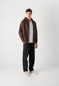 Brown zip-up hoodie with a large front pocket, worn over a white ribbed shirt, paired with loose dark jeans and beige sneakers.