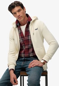 Creme zip-up hoodie med snøre, lagt over en rød plaid skjorte. Blå jeans, siddende på en skammel, med et ur på venstre håndled.