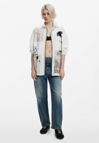 Veste blanche surdimensionnée avec des patches en mixed media, jeans bleus déchirés et baskets noires avec des lacets verts. Cheveux courts et texturés, blonds.