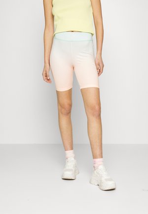 Person trägt pastellgelbes ärmelloses Oberteil, leichte Ombre-Radlerhose, rosa Socken und weiße klobige Sneakers, steht vor einem schlichten Hintergrund.
