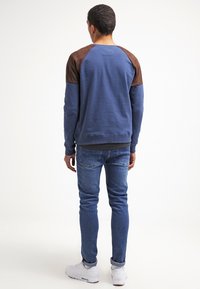 Felpa blu con accenti marroni sulle spalle, scollatura rotonda e polsini a coste. Abbinata a jeans azzurri e sneakers bianche.
