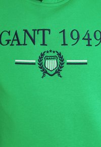 Tissu vert avec texte bleu marine "GANT 1949" au-dessus d'un blason avec des étoiles, des rayures et des branches de laurier, encadré par deux lignes horizontales.
