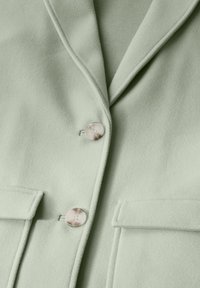 Blazer verde chiaro in morbido tessuto, con revers a punta, due tasche a toppa frontali e bottoni lisci e arrotondati.