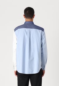 Camicia a bottoni con pannelli azzurri e blu scuro, dalla texture liscia e maniche lunghe. Presenta un orlo curvo e una piega sul retro.