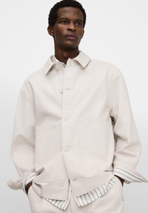 Uomo con giacca beige chiaro a bottoni indossata sopra una camicia a righe, in piedi con le mani nelle tasche contro uno sfondo uniforme.