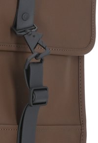 Material marrón de textura suave con una hebilla de plástico negra y correa ajustable. Presenta líneas limpias y costuras en contraste.