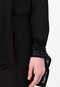 Melna blūze ar garām, plūstošām piedurknēm, kurām ir teksturēti aproces un kontrastējošs logo detaļa. Gluda auduma struktūra ar elegantu izskatu.