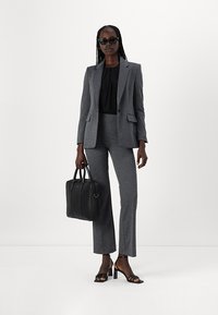 Completo grigio su misura con giacca aderente e pantaloni svasati, abbinato a una blusa nera e tacchi. Una borsa nera completa l'outfit.