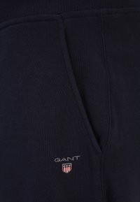 Mörk marinblå sweatshirtmaterial med ribbad mudd, har en sidficka och en liten broderad logotyp med röda och vita accenter.