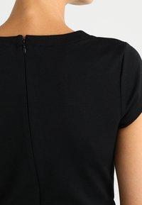 Czarny top z krótkim rękawem i okrągłym dekoltem, z dyskretnym zamkiem błyskawicznym z tyłu. Gładki materiał i dopasowany krój podkreślają design.