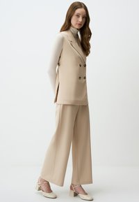 Beige dubbelzijdige vest met bruine knopen, gecombineerd met een broek met wijde pijpen. Soepele stof met een hoge hals top en beige hakken.