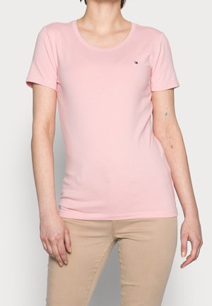 Persona con una camiseta rosa claro lisa de manga corta y pantalones beige con un pequeño logo bordado en el pecho izquierdo.