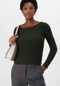 Pull à manches longues côtelé vert foncé avec un large col, porté avec un pantalon gris à motifs et tenant un sac à main gris clair.