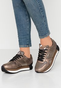 Brown athletic shoes with mesh and suede texture, black laces, white and brown sole, and subtle stripes on the tongue.