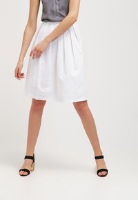 Kvinna som bär en vit plisserad kjol, en grå ärmlös topp och svarta höga klacksandaler med guldklackar, stående med benen isär.