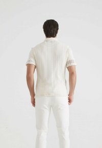 Homme se tenant de dos, portant une chemise en tricot blanche à manches courtes avec des motifs et un pantalon blanc, sur un fond uni.