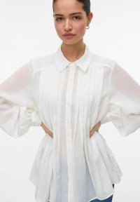 Camicia bianca con bottoni realizzata in tessuto leggero, caratterizzata da pieghe, collo a punta e maniche voluminose. Texture liscia con vestibilità rilassata.